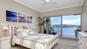 A bedroom with a bed, nightstands, a dresser, and a large window with a view of a body of water and boats. - The Commodore Penthouse (Palm Beach)