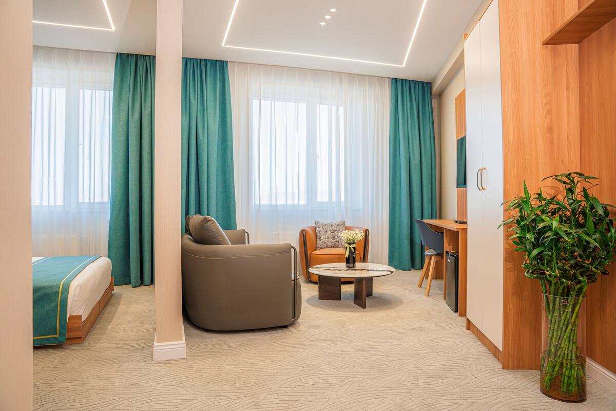 A modern hotel room with a bed, sofa, armchair, desk, and a large window with curtains.