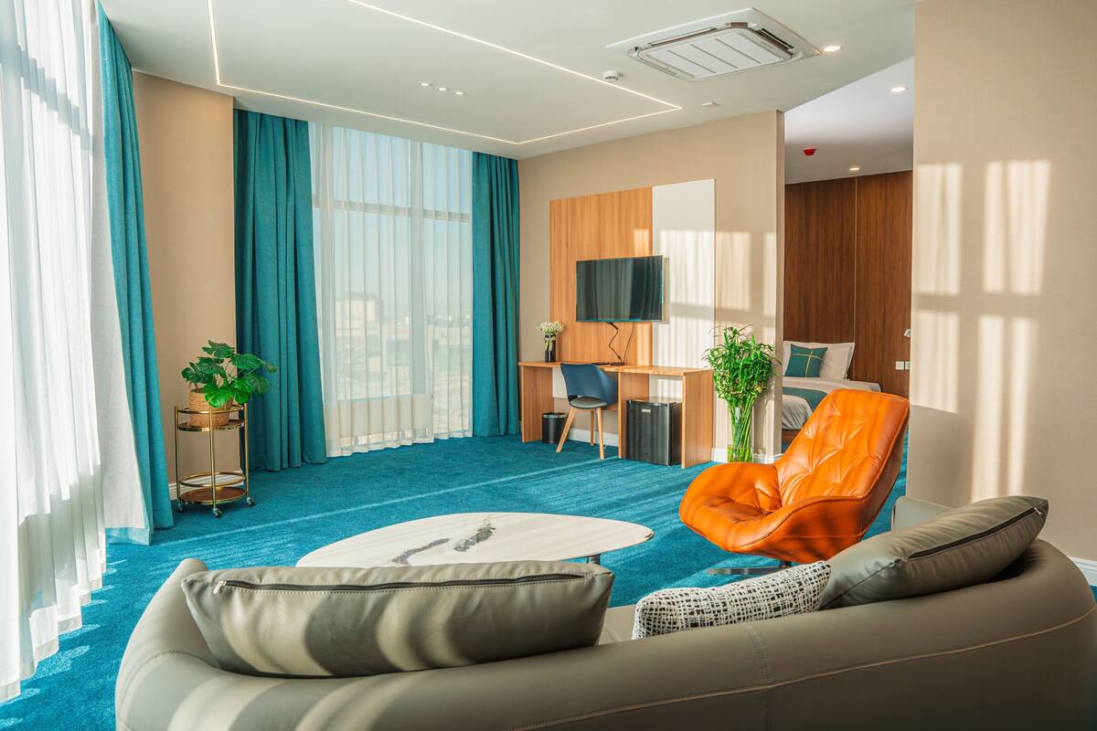 A modern hotel room with a large window, a blue carpet, a sofa, an orange chair, a desk, and a television.