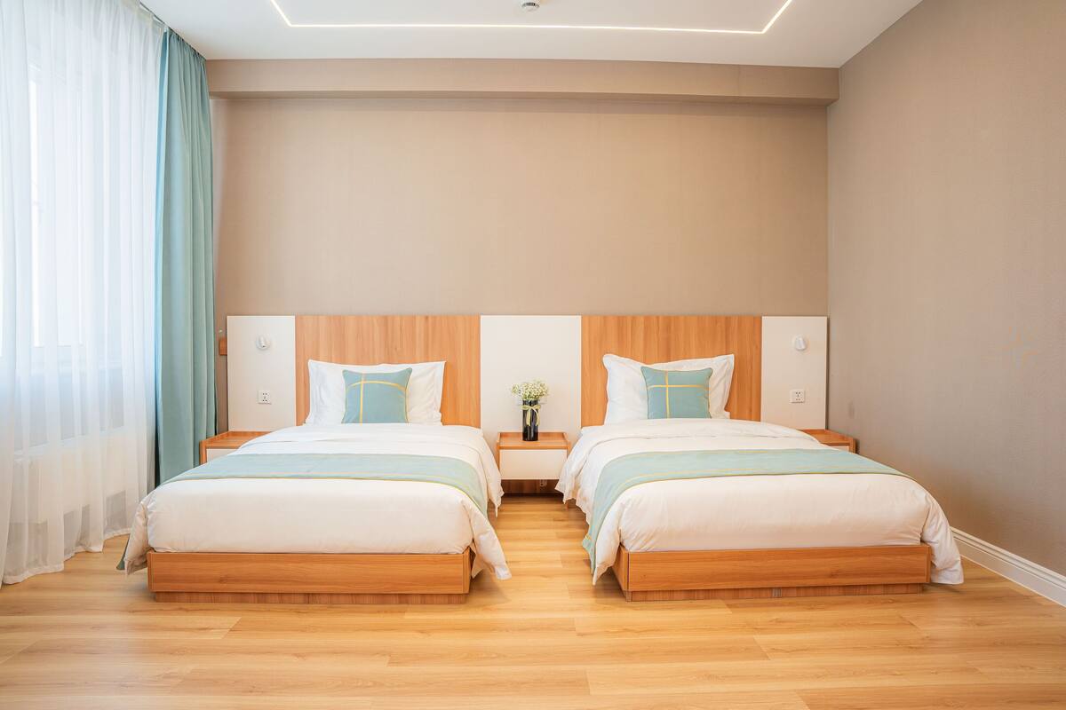A double room with two beds, wooden headboards, a bedside table with a vase, and a large window with sheer curtains.