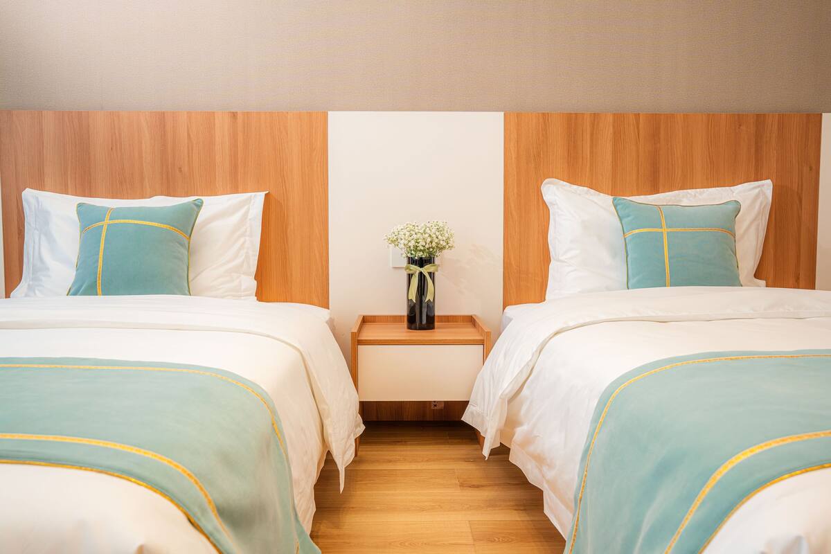 A hotel room with two beds, each with a white and teal bedspread, a wooden headboard, and a small bedside table with a vase of flowers.