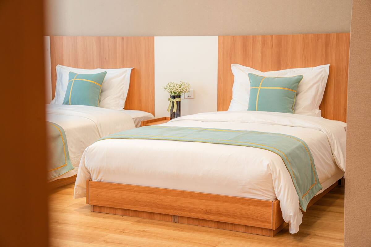 A hotel room with two beds, wooden headboards, a nightstand with a vase of flowers, and a wooden floor.
