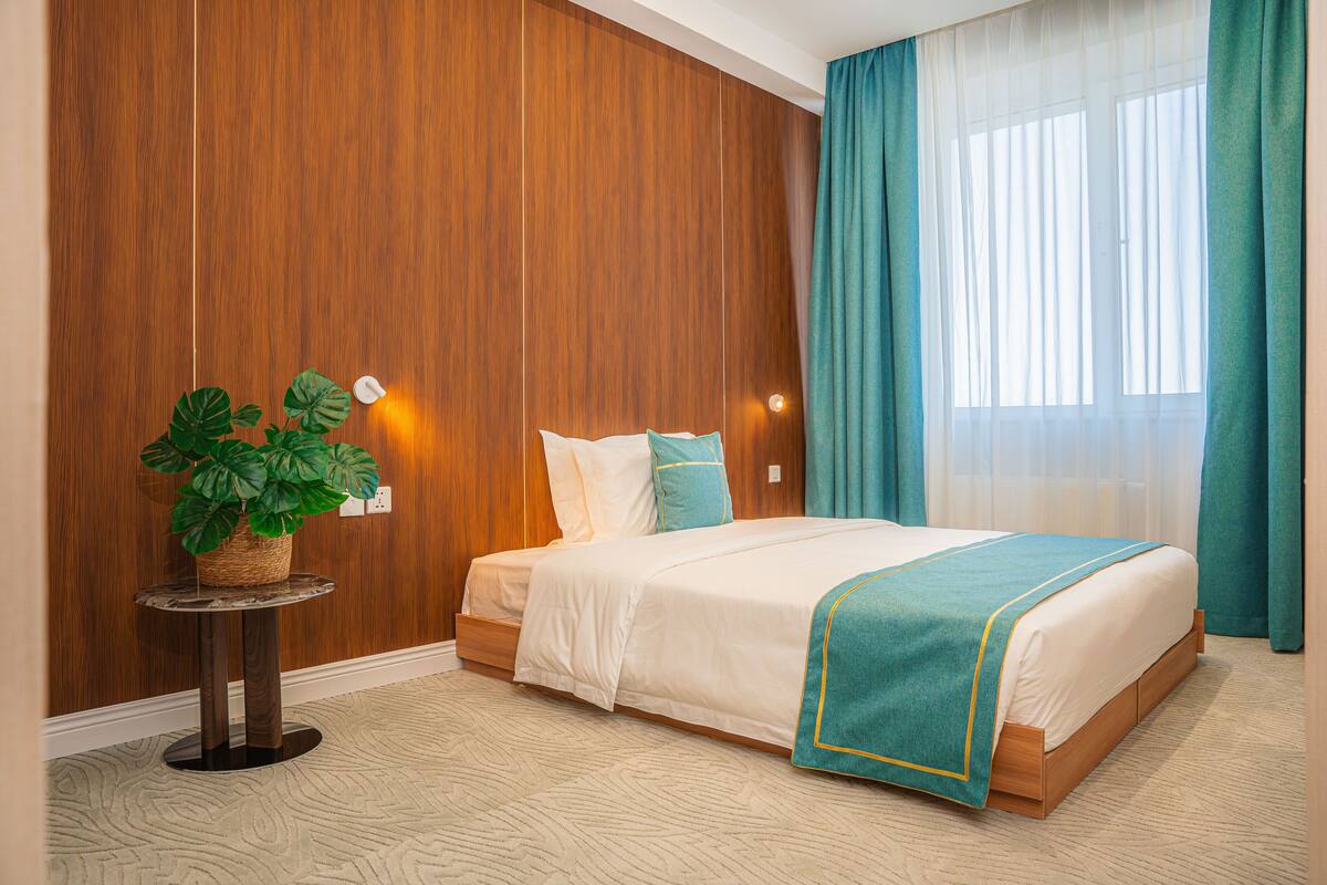 A hotel room with a wooden panel wall, a bed with white bedding and teal accents, a potted plant on a round table, and a large window with teal curtains.