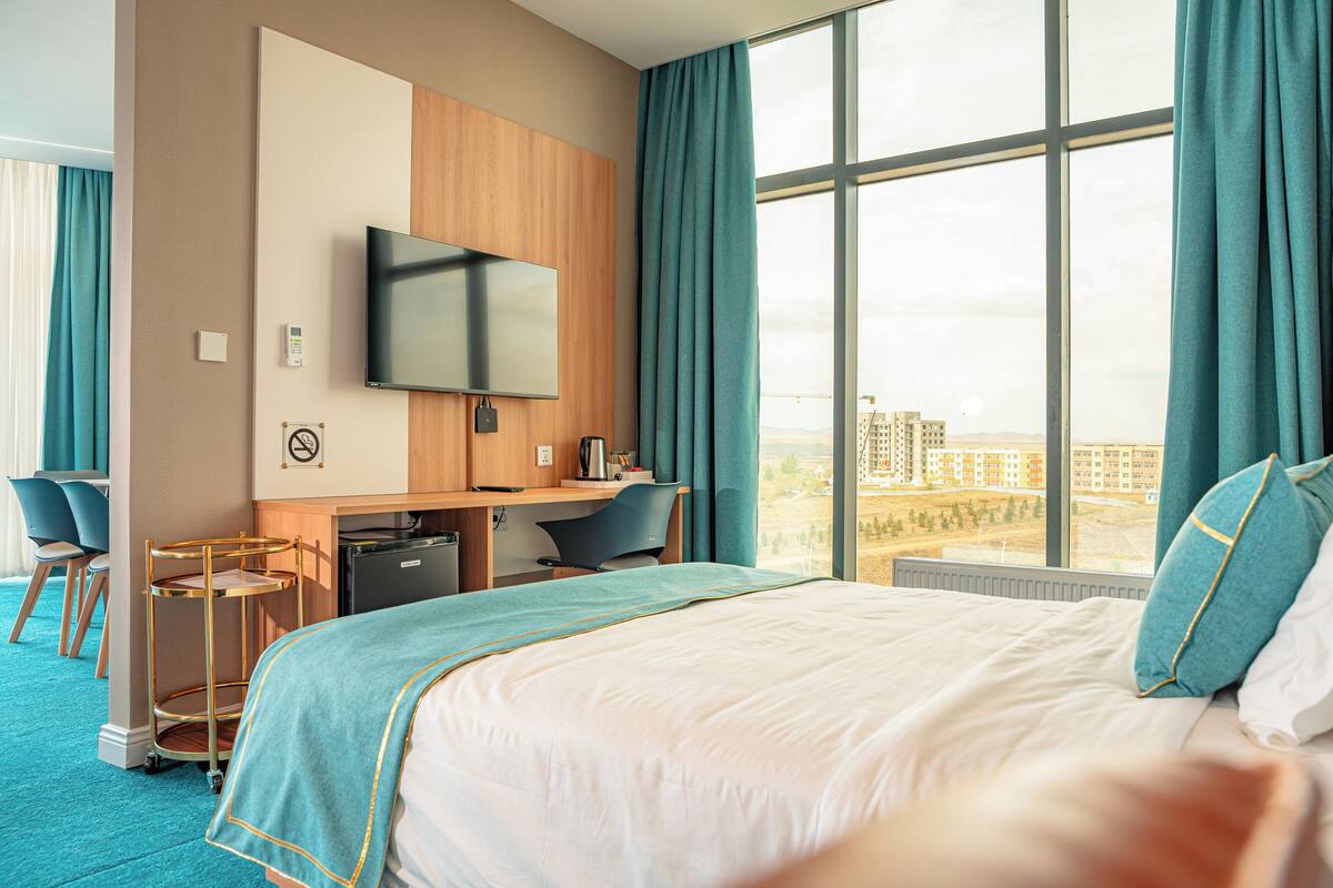 A hotel room with a large bed, a desk with a chair, a television, and a view of buildings through the window.