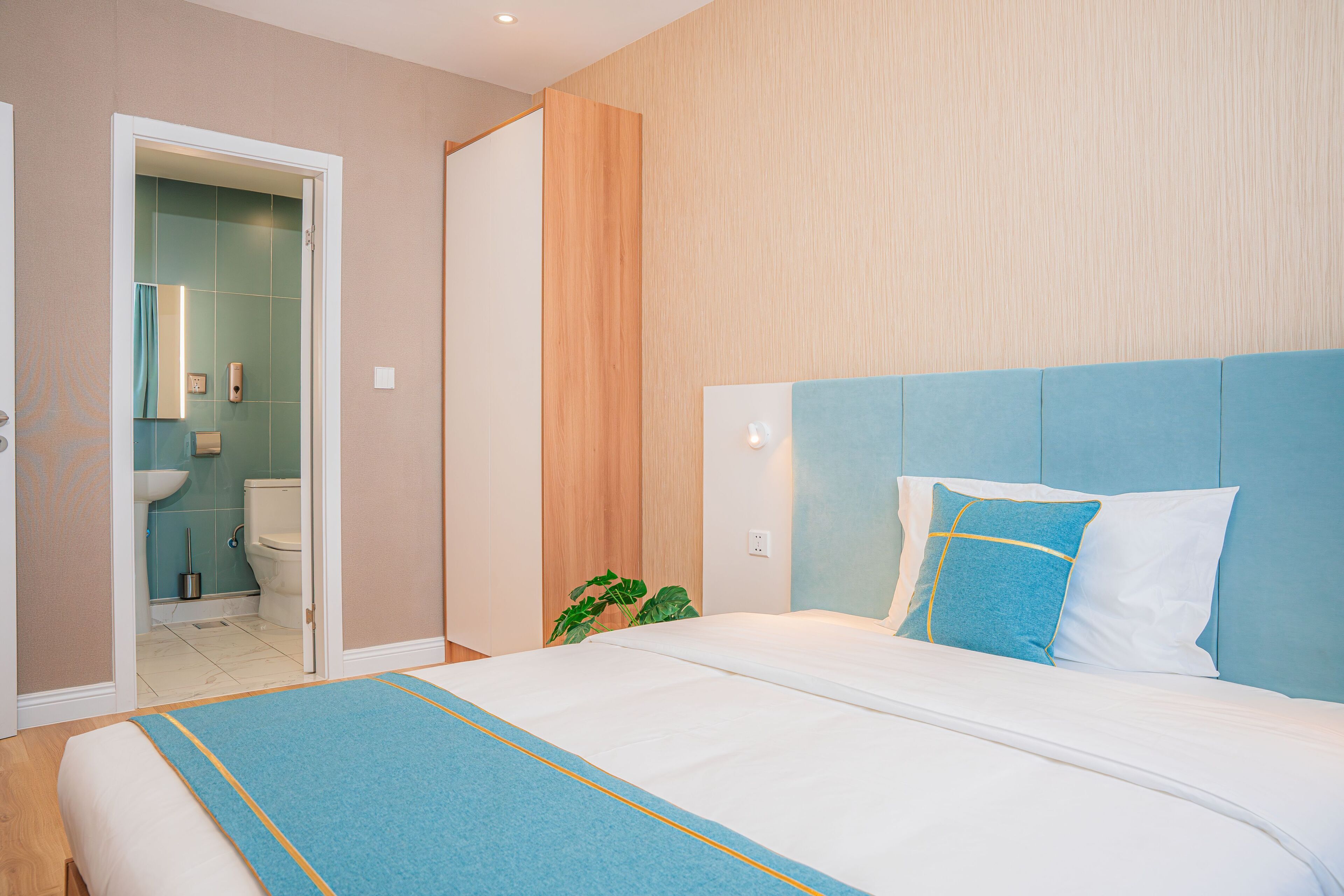 A bedroom with a bed, a bathroom visible through an open door, and a plant near the bed.
