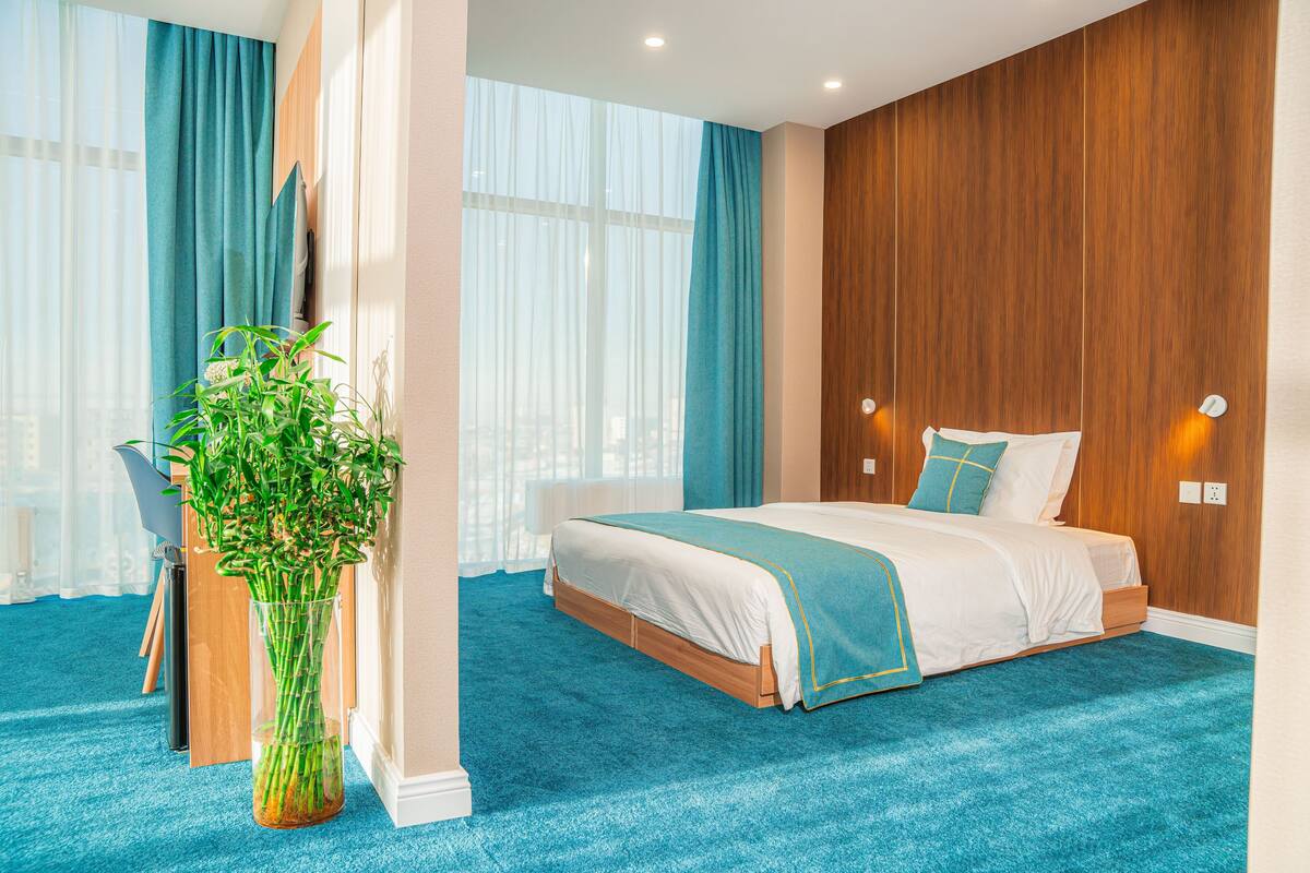 A modern hotel room with a large bed, a wooden accent wall, a blue carpet, and a potted plant.