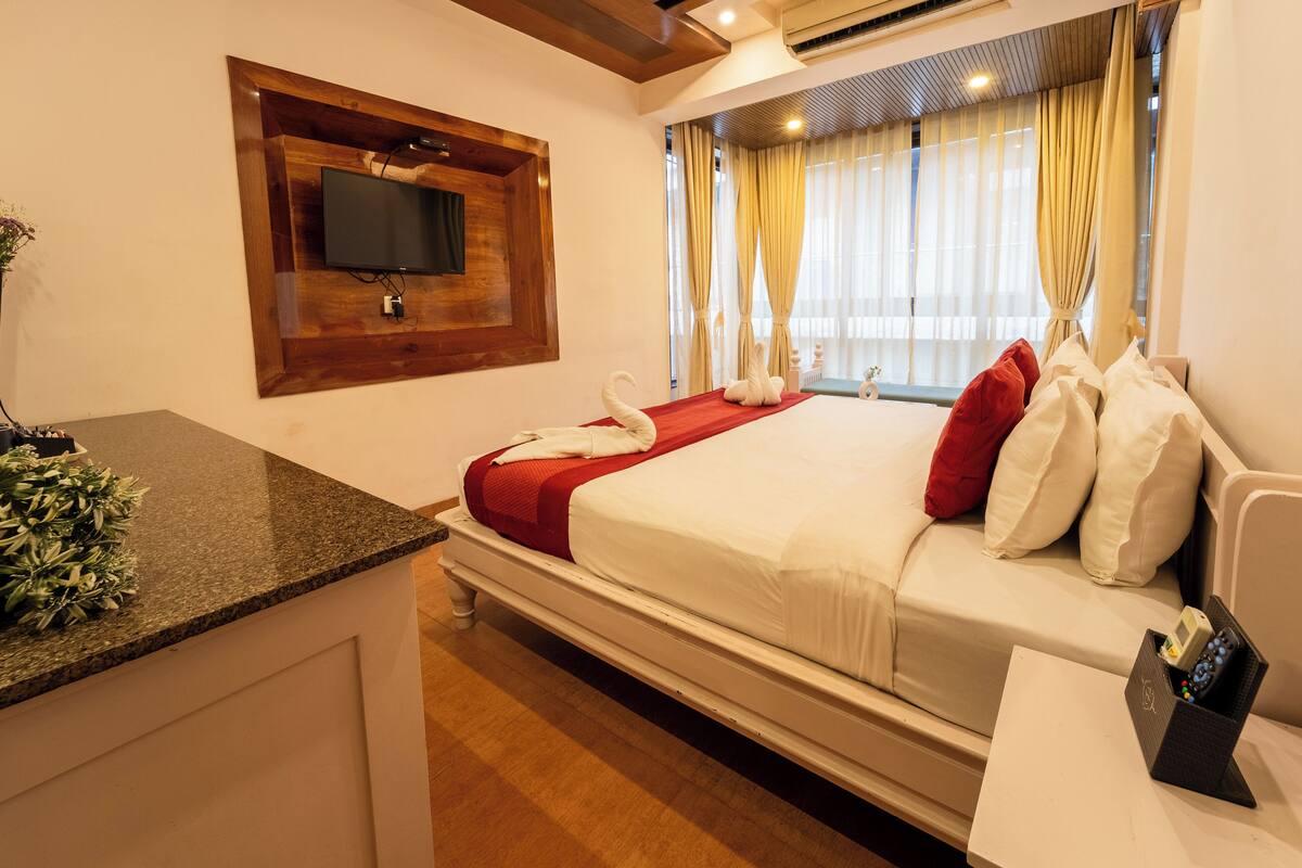 A hotel room with a bed, a TV mounted on a wooden panel, a small table with a remote control, and a window with curtains.
