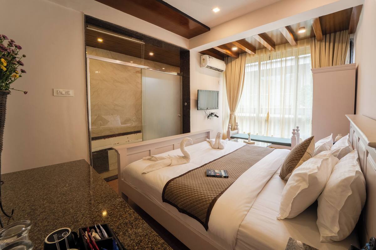 A hotel room with a large bed, a shower area, a mounted TV, and a window with curtains.