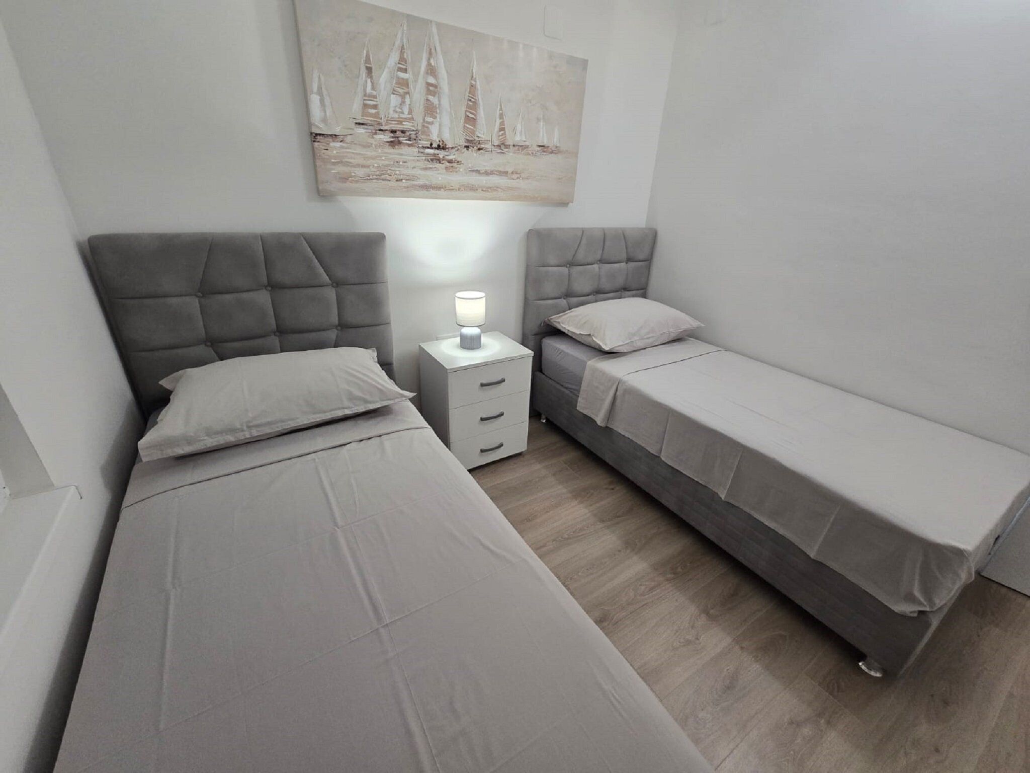 A small, modern bedroom with two single beds, a nightstand, and a wall-mounted painting.