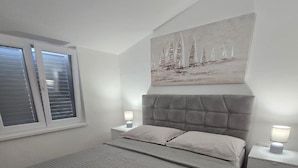 A bedroom with a large bed, two bedside tables, a window with blinds, and a painting on the wall. - Apartments Ivankovic (Nin)