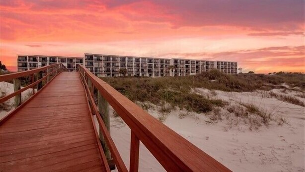 A wooden boardwalk leading to a modern building with multiple balconies, situated in a sandy area with dunes in the background. - Beacher's Lodge Studio Suite (not directly oceanfront) (Saint Augustine)