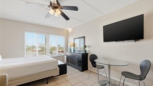 A bedroom with a bed, a dresser, a desk with two chairs, a ceiling fan, and a TV mounted on the wall. - Beacher's Lodge Studio Suite (not directly oceanfront) (Saint Augustine)
