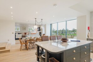 Une cuisine moderne dotée d’un grand îlot central, d’une table à manger en bois et de chaises. Des luminaires suspendus sont placés au-dessus de l’îlot, et de grandes fenêtres offrent une vue sur l’extérieur.