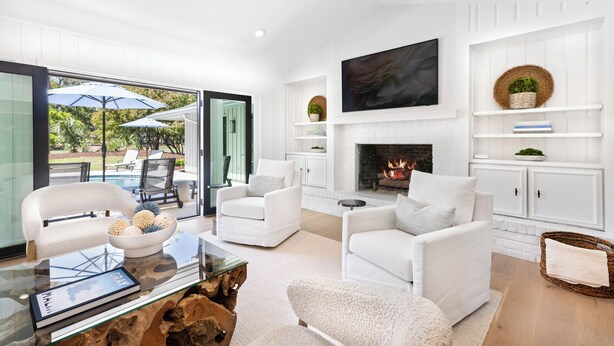 A modern living room with white furniture, a fireplace, and a glass door leading to an outdoor area with a pool and umbrellas. - 12 Woodbine Place: BESTNEST/Luxe, Private Pool, Stunning Golf View (Hilton Head Island)