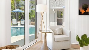A modern living room with a view of a swimming pool, a white armchair, a floor lamp, and a framed picture on the wall. - 12 Woodbine Place: BESTNEST/Luxe, Private Pool, Stunning Golf View (Hilton Head Island)