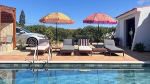 A swimming pool with lounge chairs and umbrellas, a car, and a building with a tiled roof.