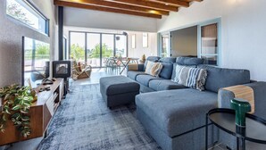 A modern living room with a large gray sofa, wooden ceiling beams, a dining area with a table and chairs, and a large window offering a view of greenery.