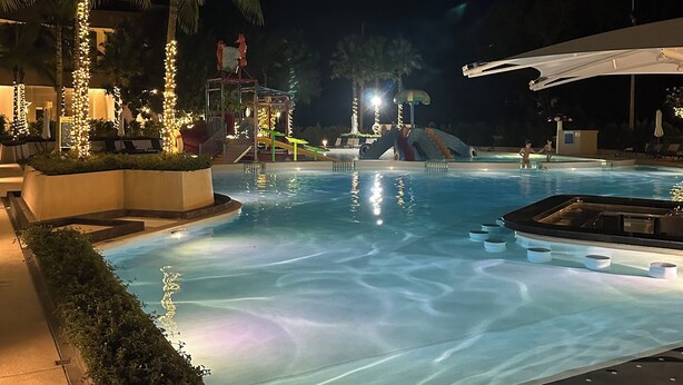 Un área de piscina nocturna con palmeras, un tobogán y una zona de estar cubierta.