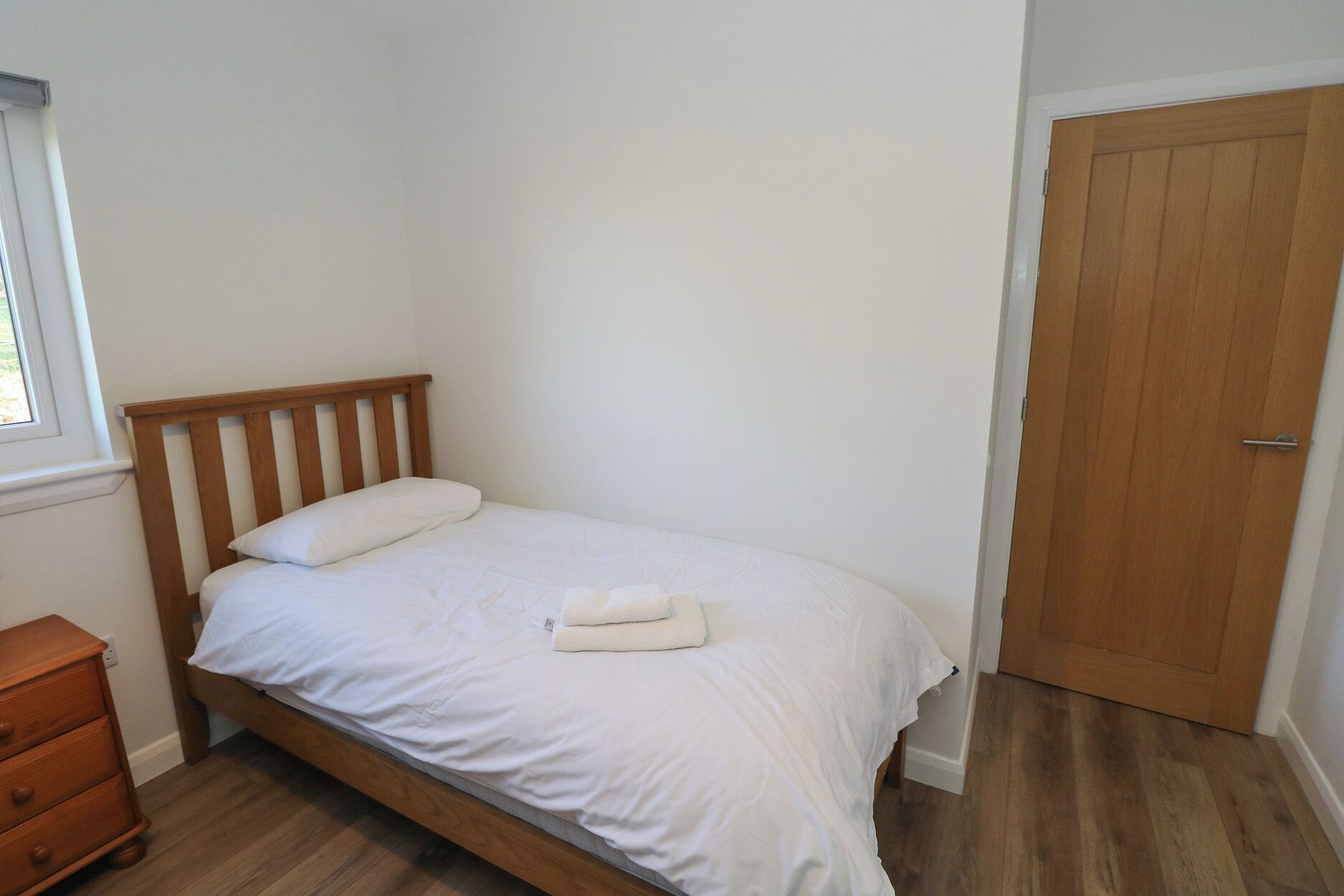 A single bed with a wooden headboard, a white mattress, and a white blanket. There is a wooden dresser to the left and a wooden door to the right.