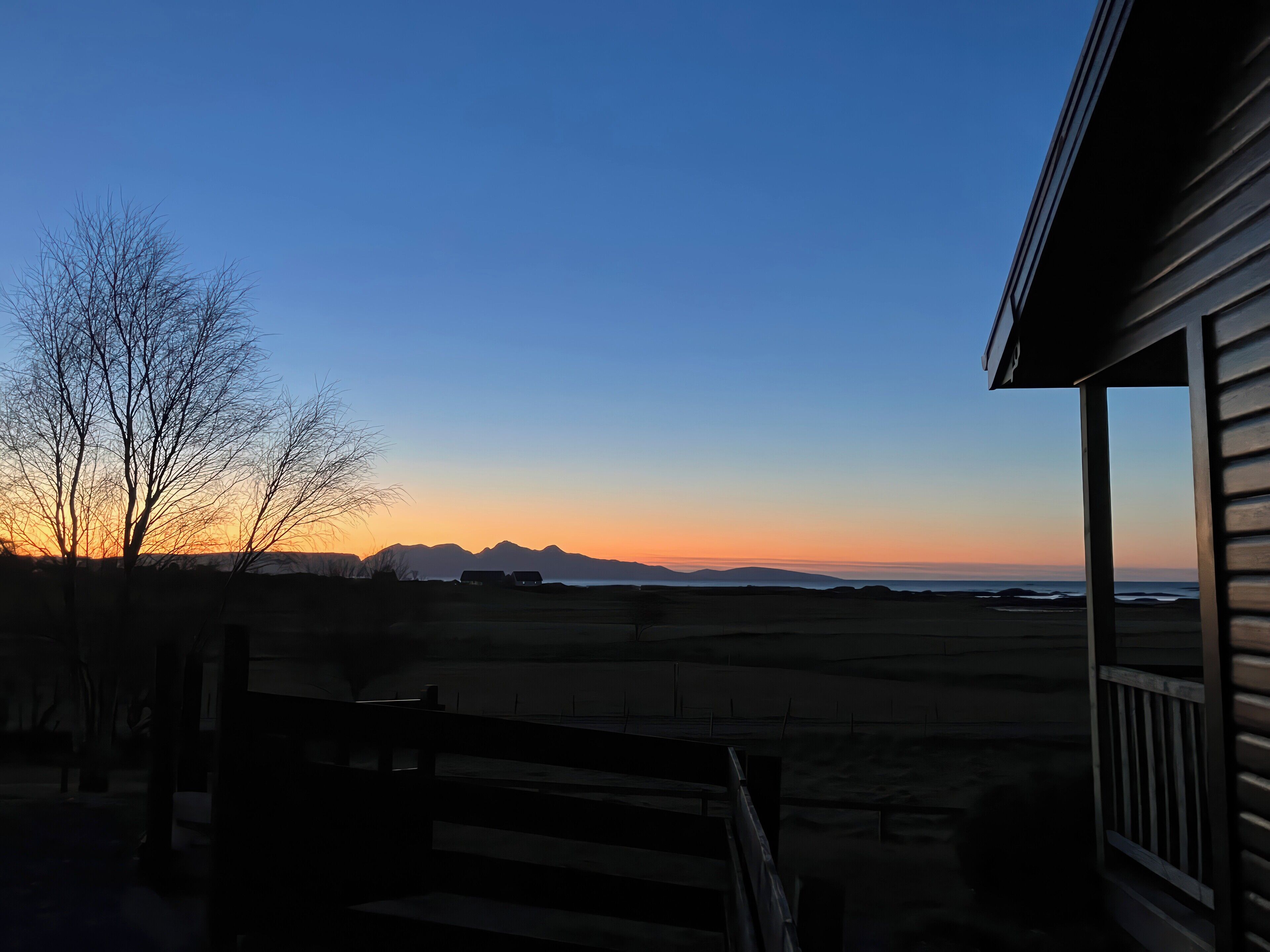 A sunset view from a porch with a wooden railing, overlooking a flat landscape and distant mountains.