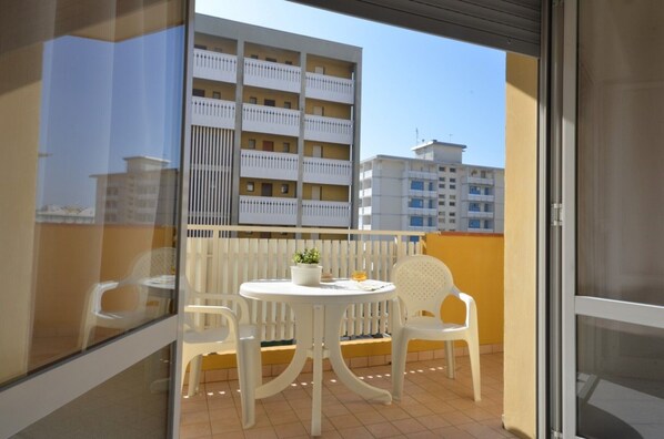 A balcony with a white table and chairs, a potted plant, and a view of a building with balconies.