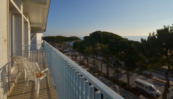 A balcony with two white plastic chairs and a small table, overlooking a street with parked cars and trees.