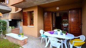 An outdoor patio with a round table and chairs, a brick planter with plants, and a grill. - Bright and cozy stay by the pool (Bibione)