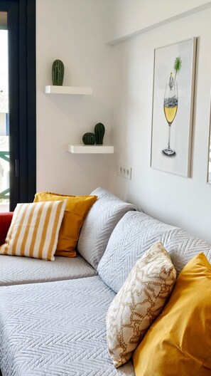 Una sala de estar moderna con un sofá gris, cojines amarillos y estampados, un cactus en un estante blanco y una obra de arte enmarcada de una copa de vino.