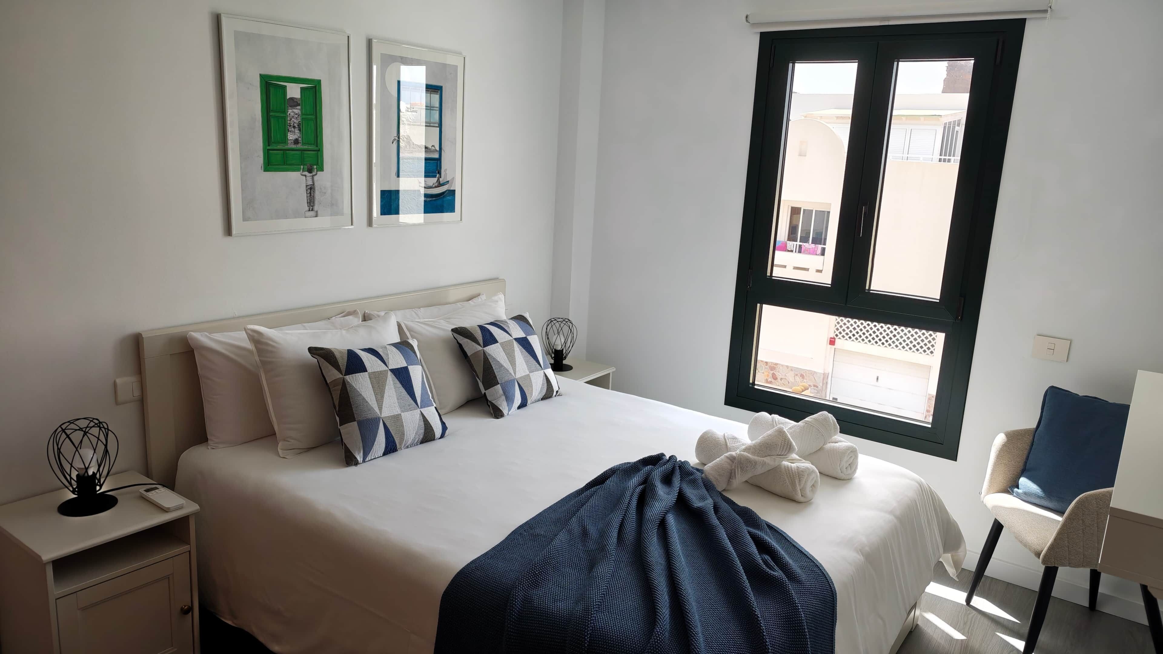 Een net opgemaakt bed met kussens, een nachtkastje met een lamp, en een raam met uitzicht op het omliggende landschap.