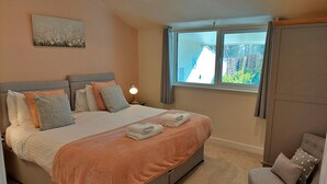Een slaapkamer met een bed, kussens, een nachtkastje met lamp, een raam met gordijnen en uitzicht op het omliggende landschap.