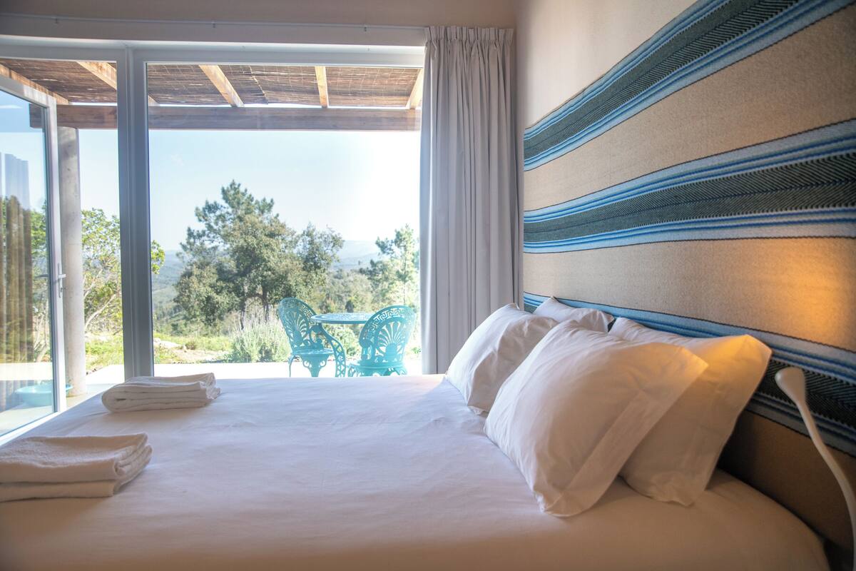 A hotel room with a bed, a view of the outdoors, and a table and chairs outside.
