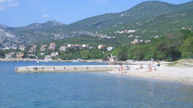 A coastal area with a sandy beach, a pier extending into the water, and buildings in the background. - Apartments Valentinovo (Novi Vinodolski)