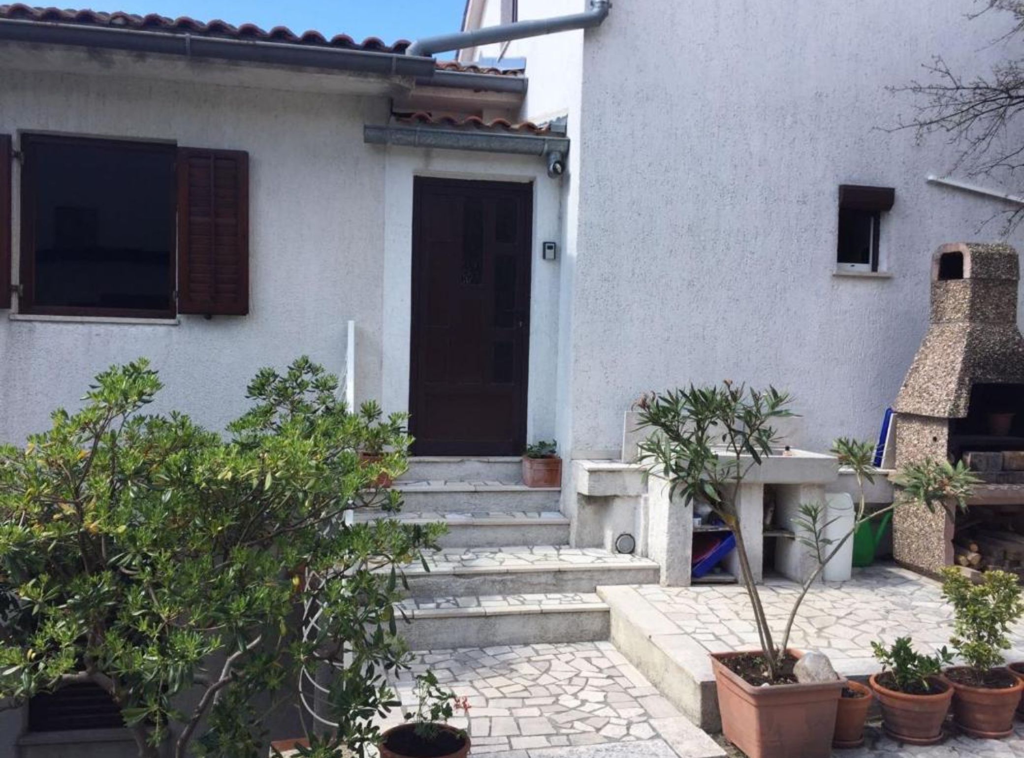 A building with a tiled roof, a front door, and a small window. There are steps leading to the door, and potted plants are placed around the entrance.