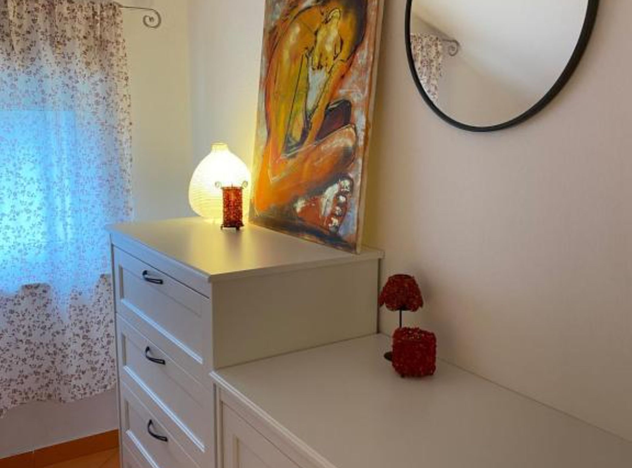 A bedroom with a bed, a dresser, a painting, a lamp, and a red decorative item.