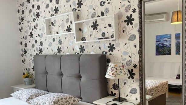 A bedroom with a grey tufted headboard, a white bedside table with a lamp, and a floral-patterned wall. - Apartments Lara Rab (Rab)