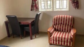A room with a bed, a dining table with chairs, and a striped armchair. - The Manor. (Lomé)