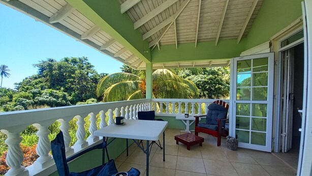 A covered balcony with a table and chairs, a view of lush greenery, and a door leading to the interior.