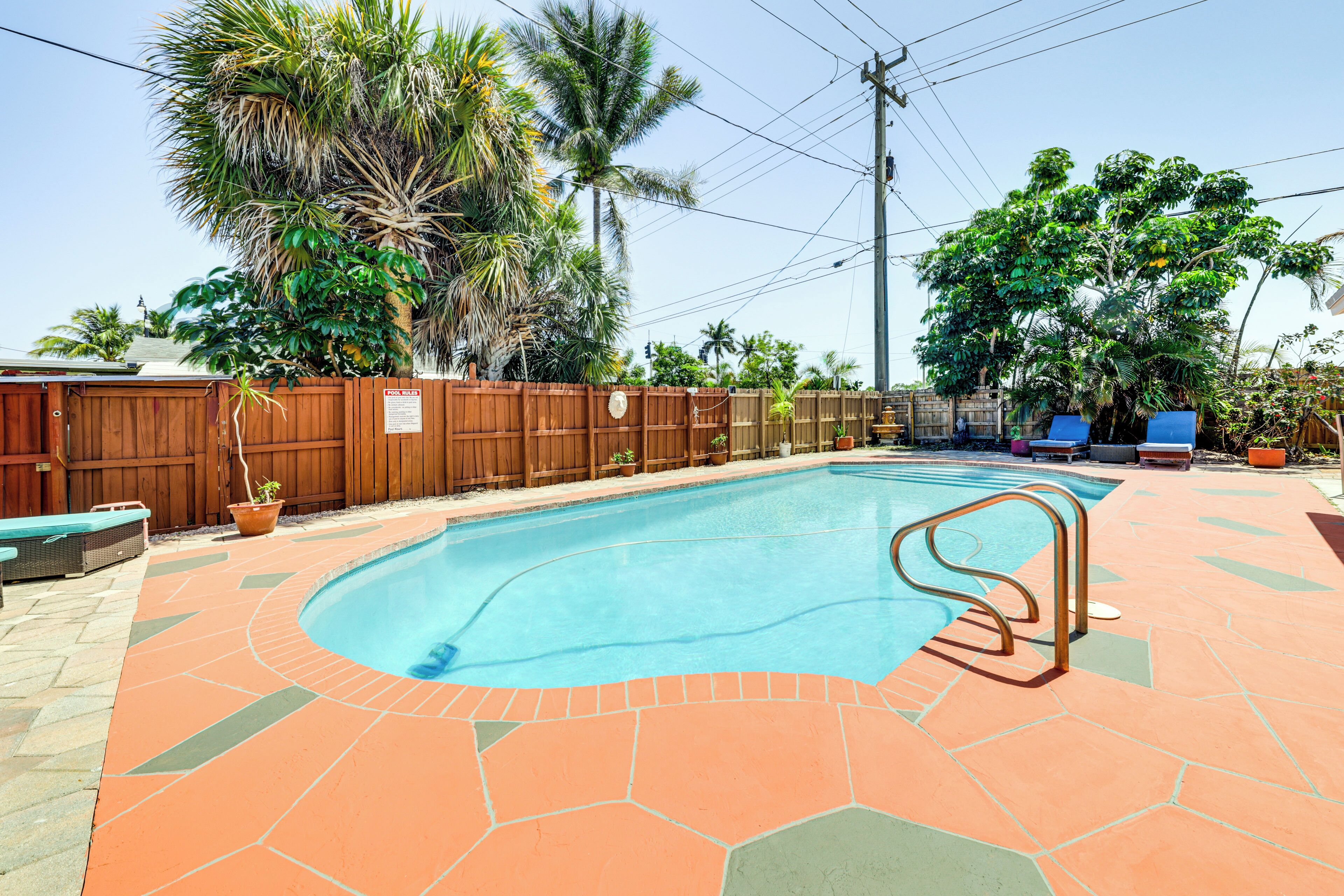 A swimming pool with a wooden fence, palm trees, and lounge chairs.