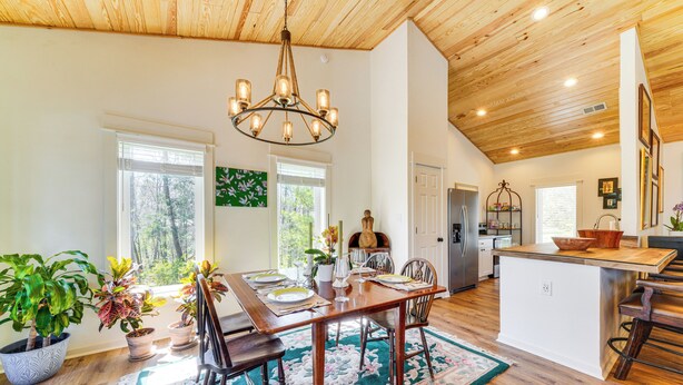 A spacious dining area with a wooden table, chairs, and a chandelier. There are potted plants and a view of the outdoors through large windows. - Hot Tub, Deck & Fire Pit: Chic Louisville Home! (Louisville)