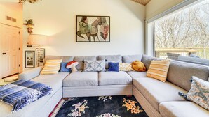 A modern living room with a grey sectional sofa, a black floral rug, and a large window with a view of trees. - Hot Tub, Deck & Fire Pit: Chic Louisville Home! (Louisville)