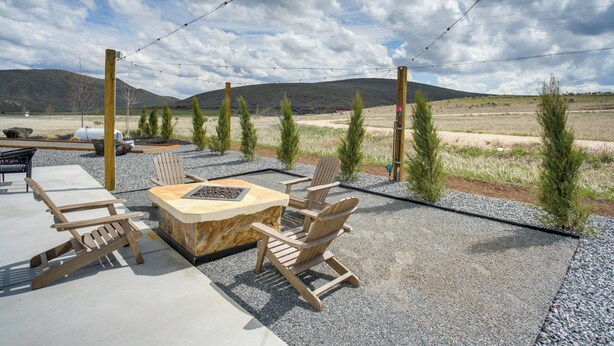An outdoor seating area with Adirondack chairs and a fire pit, surrounded by gravel and small trees, with mountains in the background.