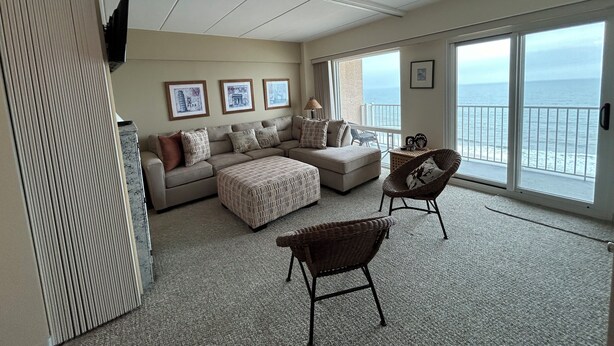A spacious living room with a large window offering a view of the ocean, a sofa set, a chair, and a flat-screen TV mounted on the wall.