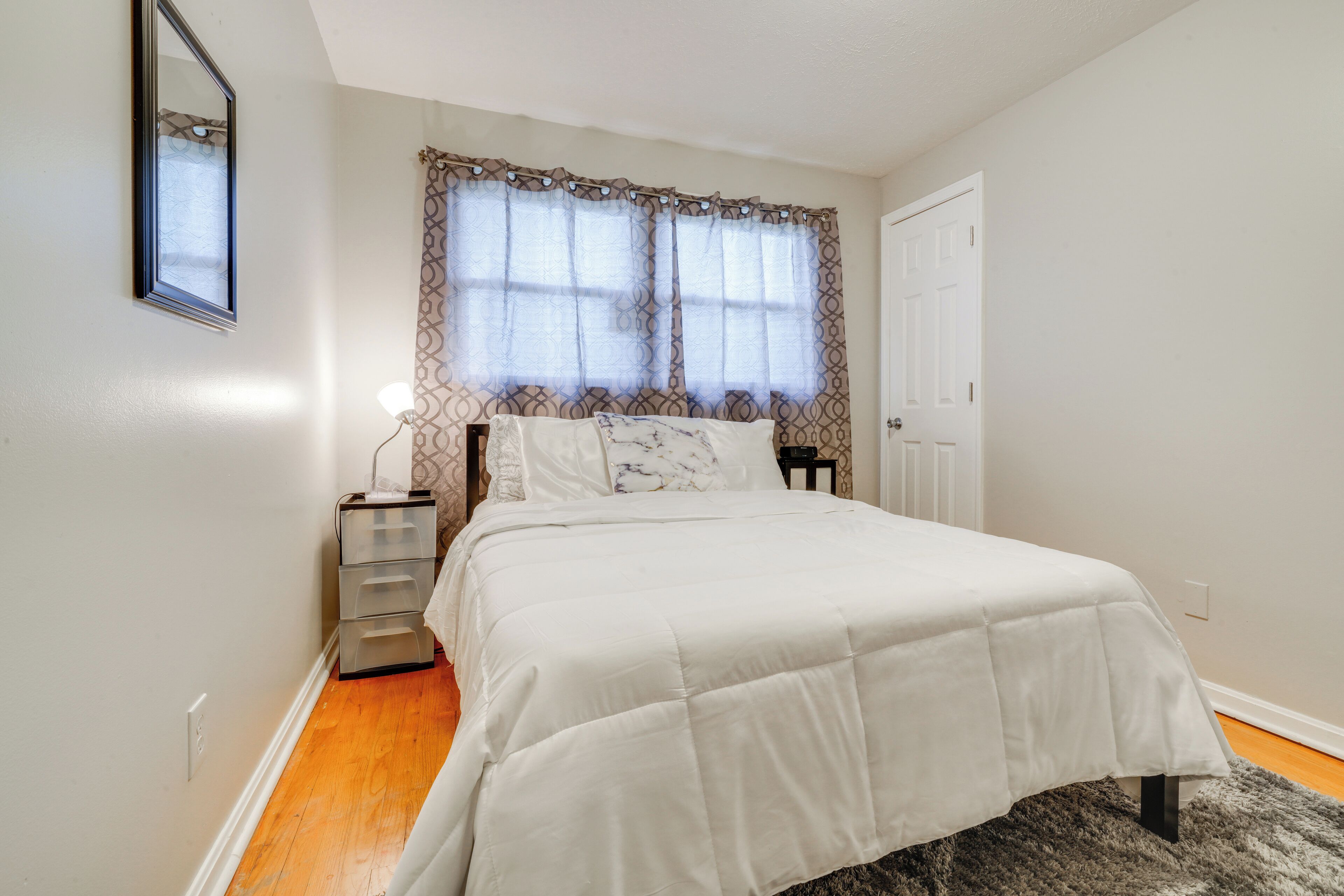 A bedroom with a bed, a nightstand, a mirror, and a window with patterned curtains.