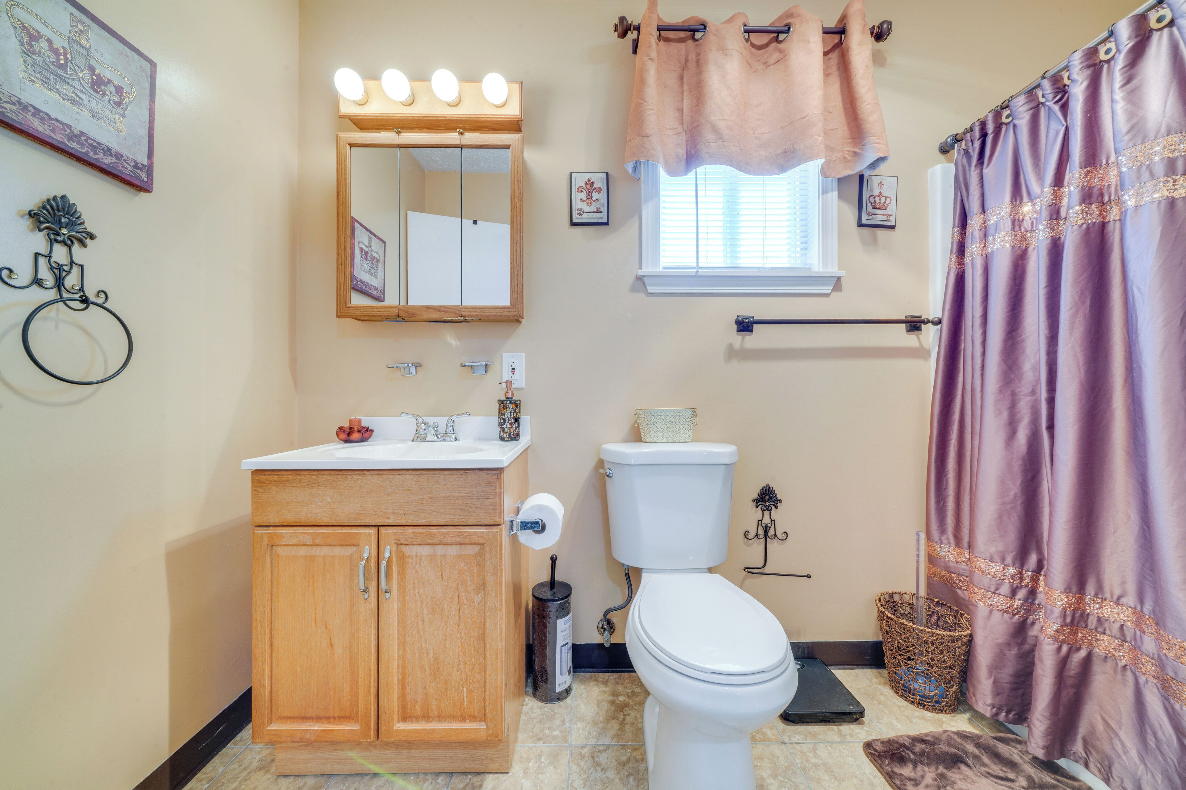 Een badkamer met een houten wastafelmeubel, toilet, spiegel met verlichting, raam met gordijn en een handdoekhouder.