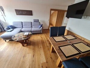 A living room with a sofa, a coffee table, a wooden dining table with placemats, and a wooden door.