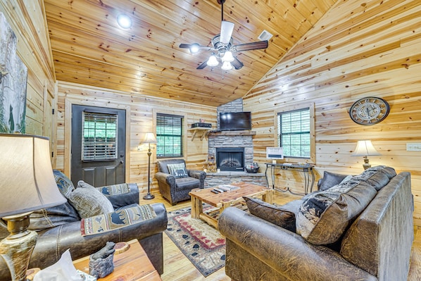 A log cabin interior with a fireplace, wooden furniture, and a ceiling fan. - Private Hot Tub & Deck: Woodsy Broken Bow Retreat (Broken Bow)