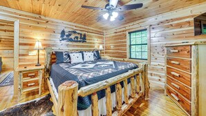 A wooden cabin bedroom with a bed, bedside lamps, a ceiling fan, and a window.