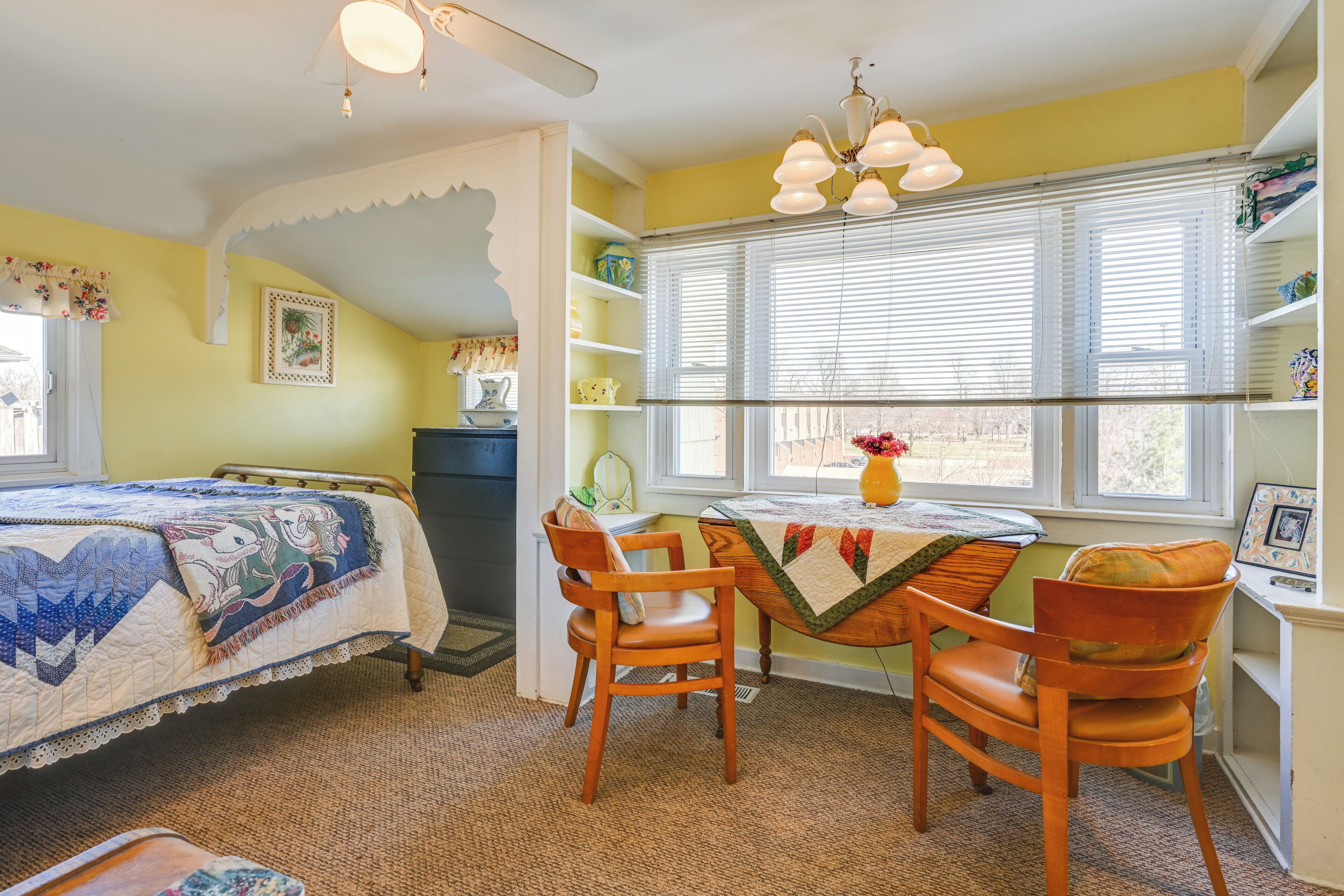 A bedroom with a bed, two wooden chairs, a table with a quilt, a window with blinds, and shelves with decorative items.