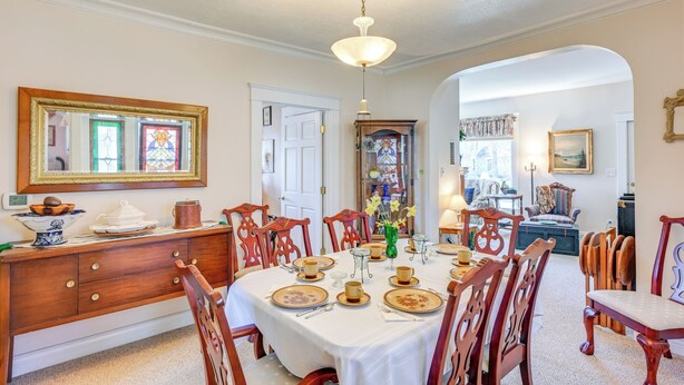 A dining room with a round table set for six, surrounded by wooden chairs. A wooden buffet unit with a mirror above it is visible.