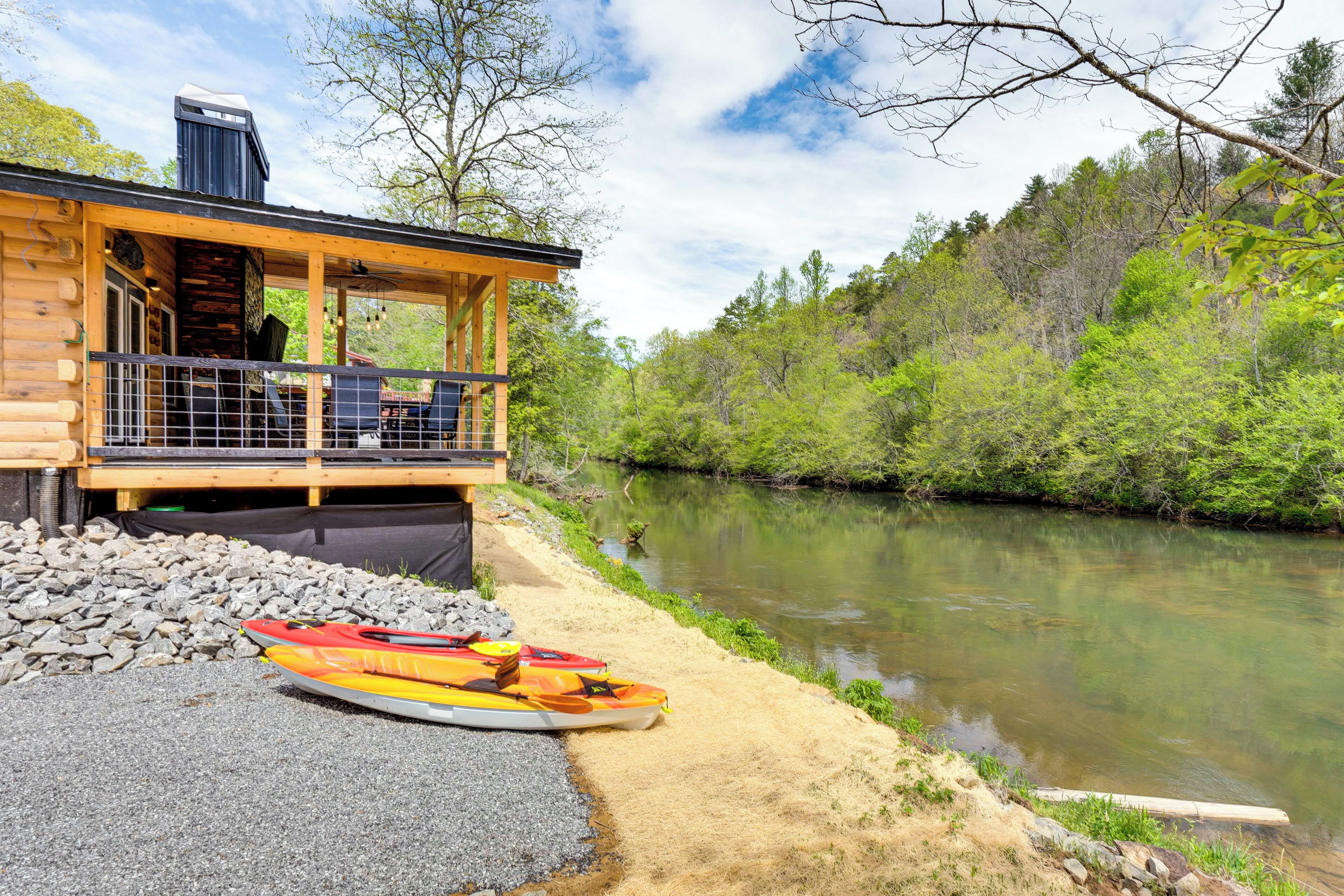 Une cabane en rondins avec une terrasse donnant sur une rivière, des kayaks sur la rive et une colline boisée en arrière-plan.
